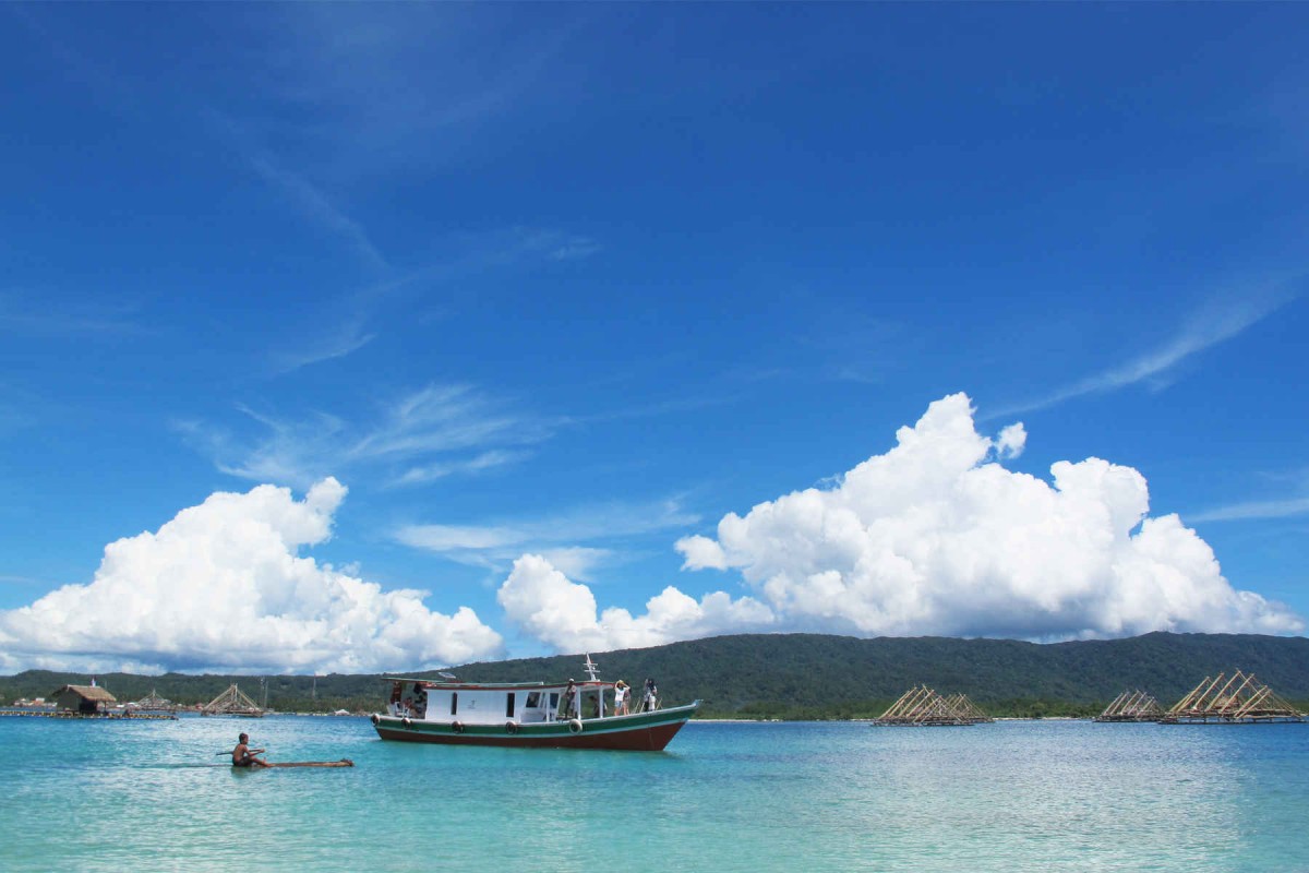 Pulau Peucang dengan langit biru dan awan putih, berpadu dengan air laut biru jernih dan perbukitan yang mengelilinginya, ada perahu nelayan yang sedang berlayar.